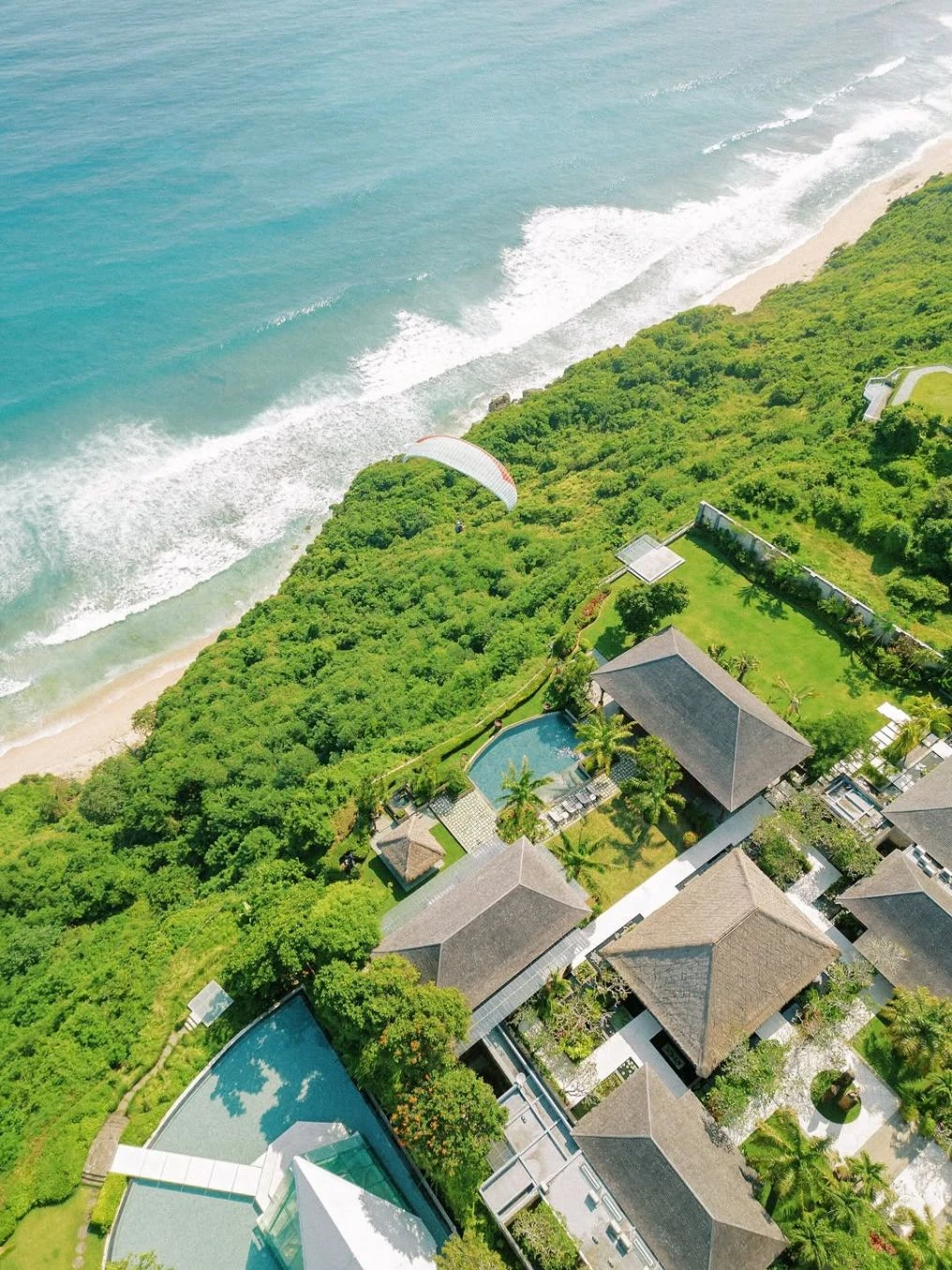 Aerial view of Tirtha Bali: thatched pavilions and pools on a green clifftop above the beach and surf